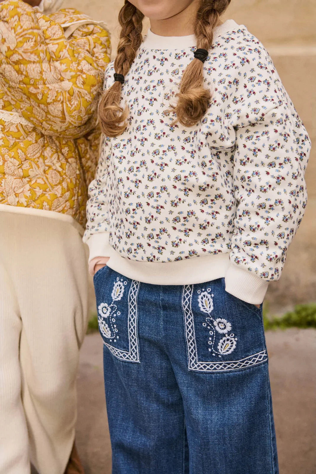 A child in a white floral sweatshirt and Louise Misha Girls' Flofi Pants with an elastic waist stands next to another child wearing a yellow patterned jacket.