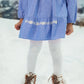 A child stands in the snow wearing the Girls' Orana Dress by Louise Misha, featuring blue fabric with white lace details, paired with white tights and brown boots. The child's face is not visible.