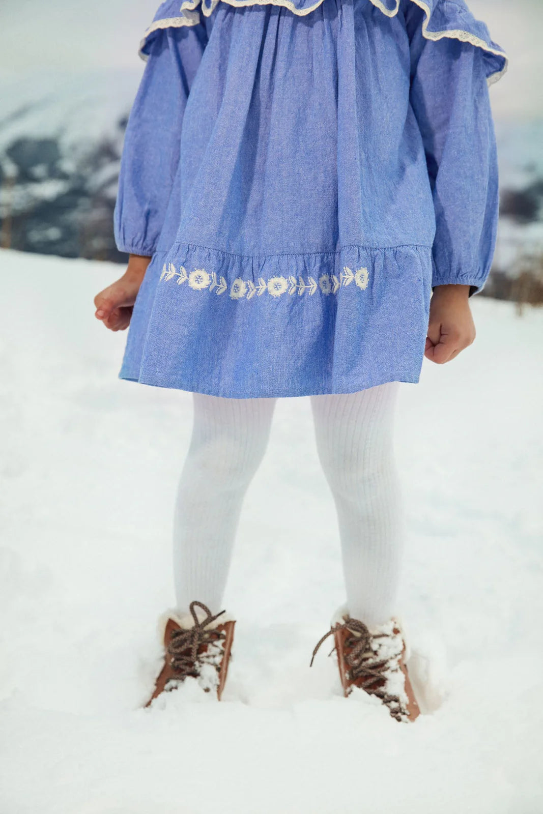 A child stands in the snow wearing the Girls' Orana Dress by Louise Misha, featuring blue fabric with white lace details, paired with white tights and brown boots. The child's face is not visible.