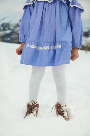 A child stands in the snow wearing the Girls' Orana Dress by Louise Misha, featuring blue fabric with white lace details, paired with white tights and brown boots. The child's face is not visible.