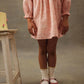A girl wears the Louise Misha Girls' Saralou Dress in pink with white socks and pink shoes, standing with crossed legs by a small white stool topped with a yellow cake against a plain background.