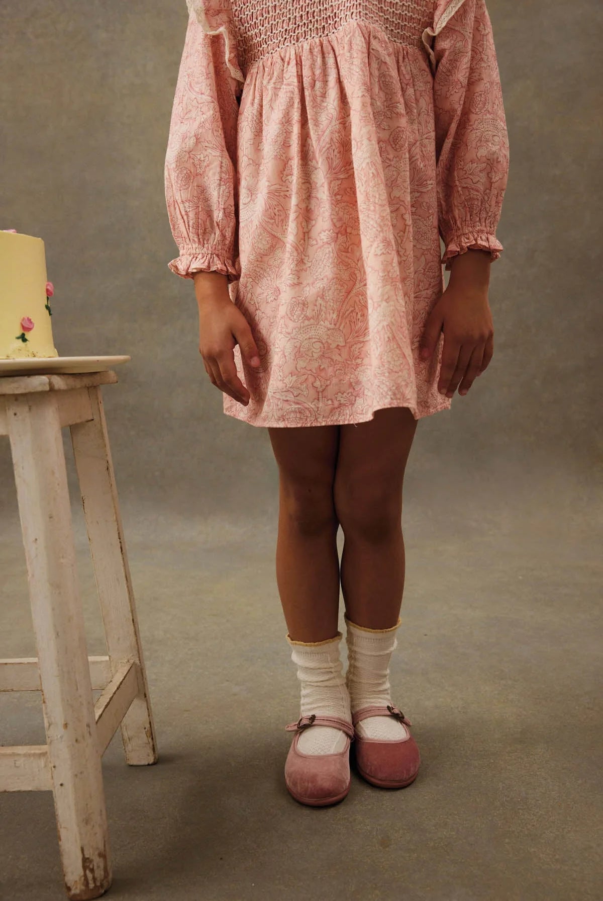 A girl wears the Louise Misha Girls' Saralou Dress in pink with white socks and pink shoes, standing with crossed legs by a small white stool topped with a yellow cake against a plain background.