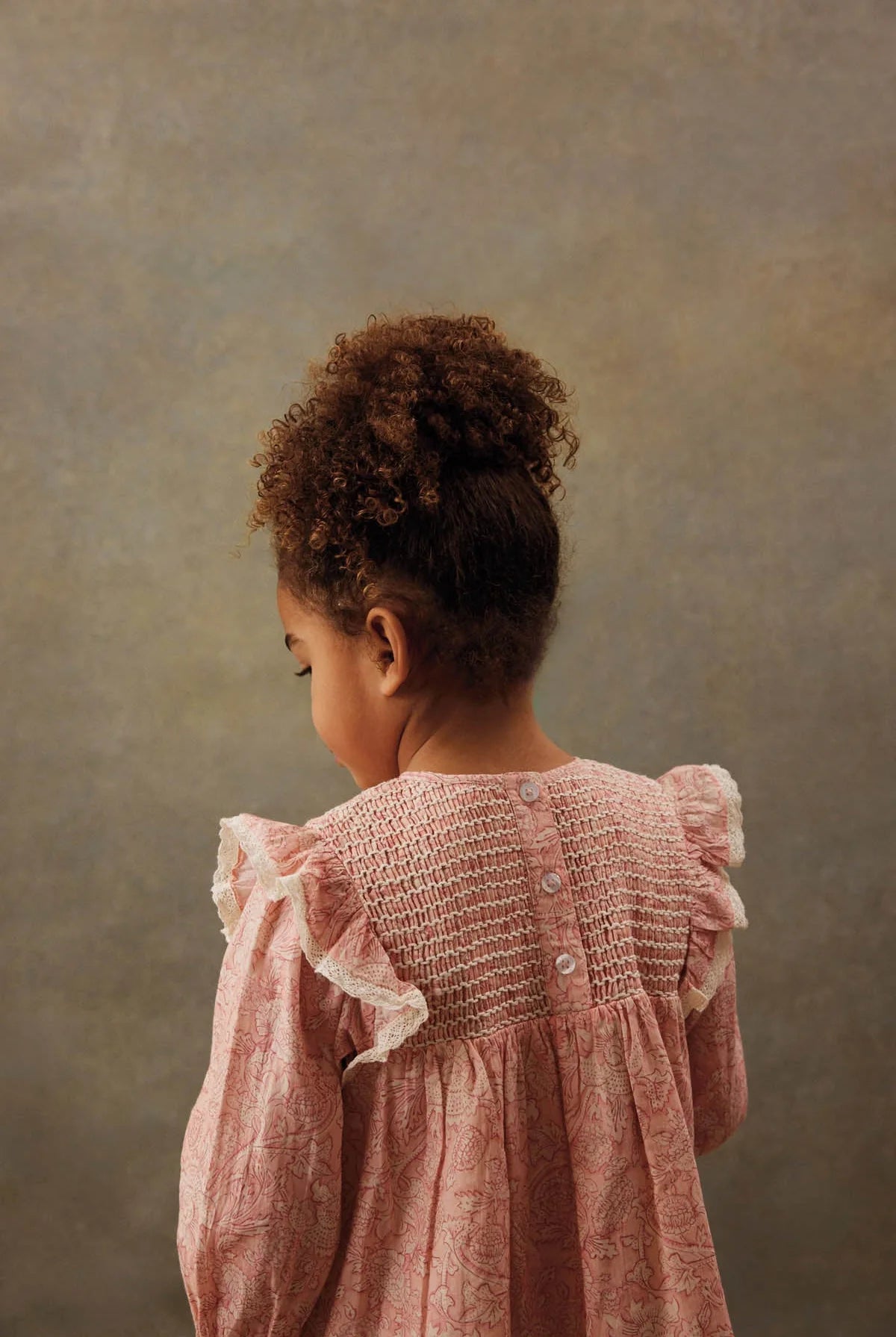 A young child with curly hair, wearing the Louise Misha Girls' Saralou Dress in pink cotton gauze with ruffled sleeves, stands facing away from the camera against a neutral background.