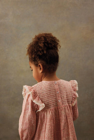 A young child with curly hair, wearing the Louise Misha Girls' Saralou Dress in pink cotton gauze with ruffled sleeves, stands facing away from the camera against a neutral background.