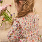 A young girl with long, wavy hair wears the Louise Misha Girls' Saralou Dress and holds a bouquet of flowers, standing by a flower-adorned wall.