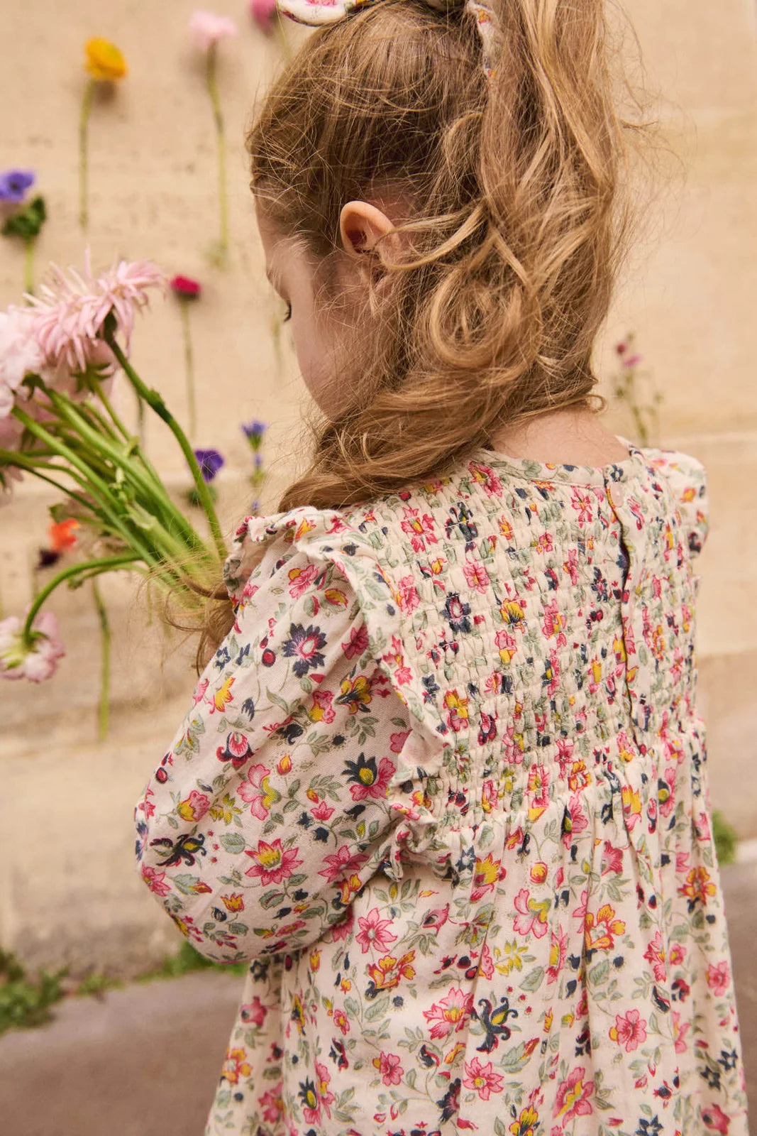 A young girl with long, wavy hair wears the Louise Misha Girls' Saralou Dress and holds a bouquet of flowers, standing by a flower-adorned wall.