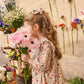 A young girl wearing the Louise Misha Girls' Saralou Dress holds a bouquet of pink flowers, standing beside a wall adorned with vibrant blooms.