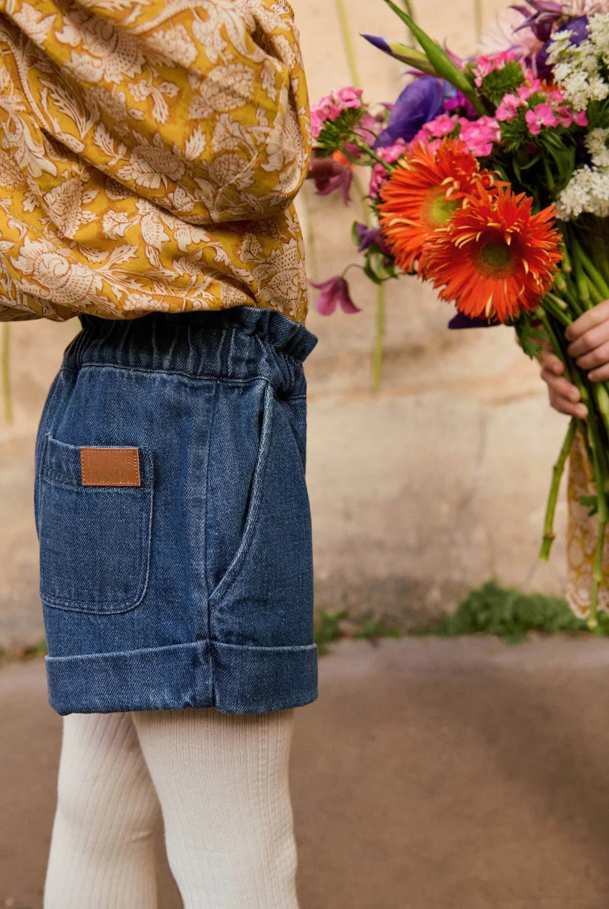 A child stands next to a person holding a bouquet of colorful flowers, wearing a patterned yellow top, white tights, and Louise Misha Girls' Virgina Shorts with a wide fit and elasticized waist.