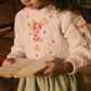 A young girl with curly hair wears a cream sweater with floral embroidery and the Louise Misha Girls' Cephee Skirt, featuring an elasticized waistband, as she holds an open book and looks down at the pages.