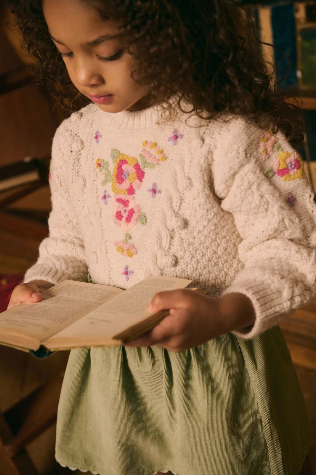 A young girl with curly hair wears a cream sweater with floral embroidery and the Louise Misha Girls' Cephee Skirt, featuring an elasticized waistband, as she holds an open book and looks down at the pages.