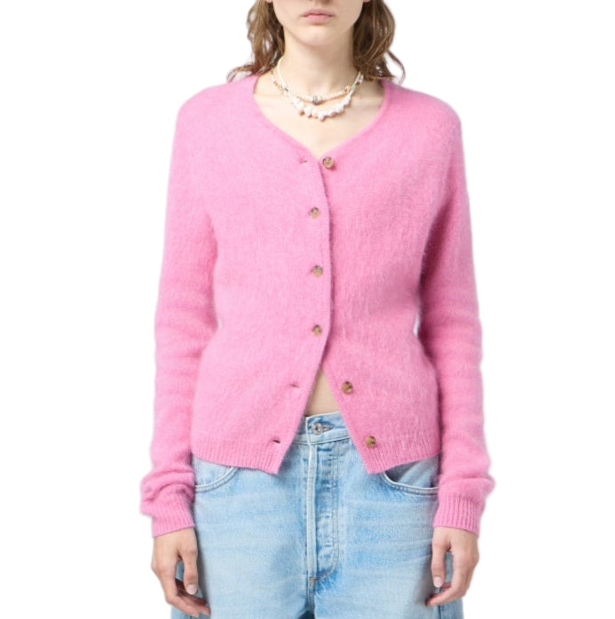 A woman wears the Grizzly Cashmere Cardigan by Guest in Residence—a pink knit button-up with a brushed finish—paired with light blue jeans and layered necklaces, standing against a white background.