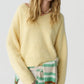 A person wearing the Grizzly Double V Sweater in soft yellow by Guest in Residence—a lightweight cashmere pullover with a double V neckline—paired with light pants and a striped garment tied around the waist, stands against a plain background.