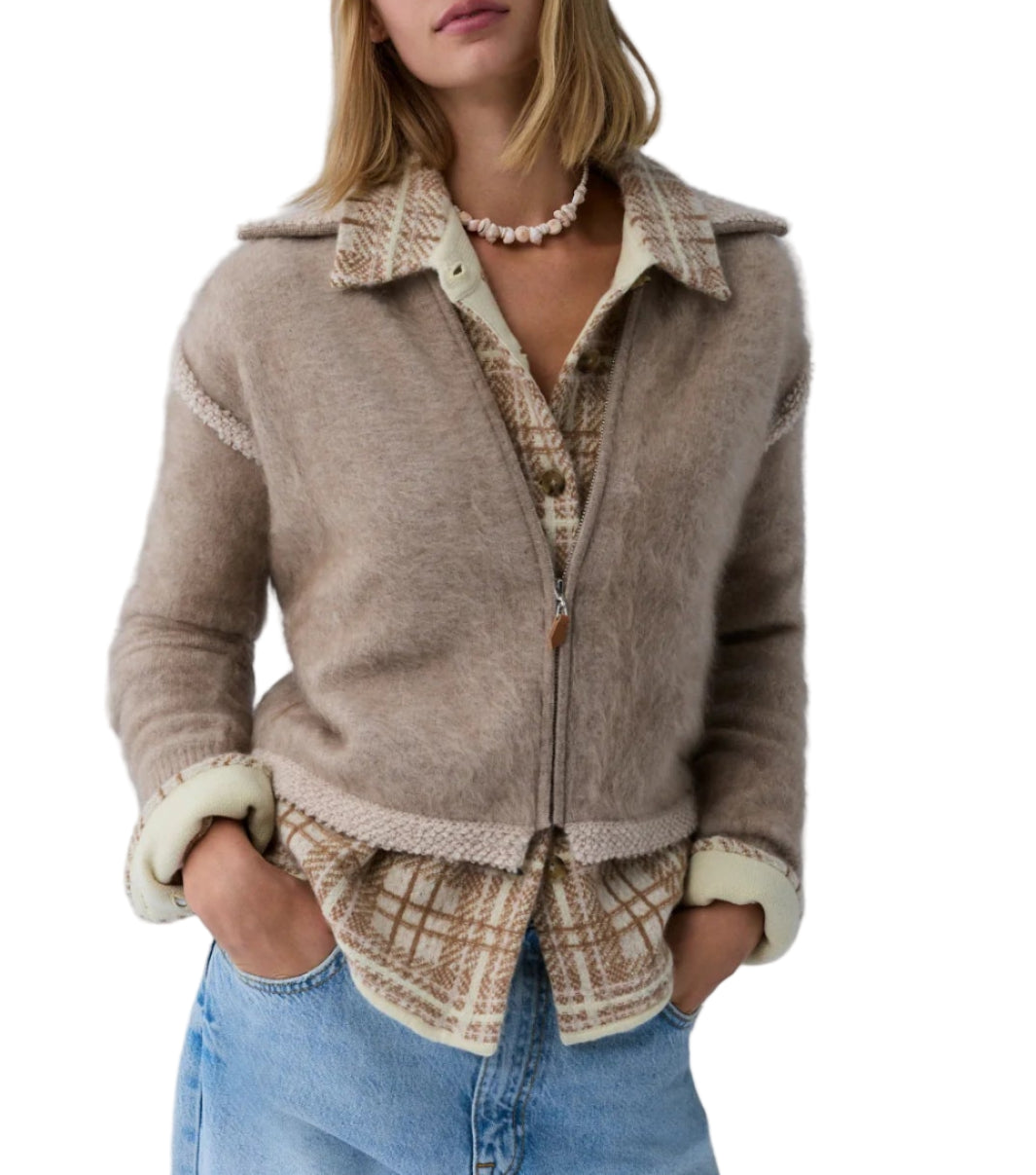 A person wears the Grizzly Frankie Jacket by Guest in Residence over a plaid shirt and blue jeans, hands in pockets. They have shoulder-length hair and accessorize with a shell necklace.