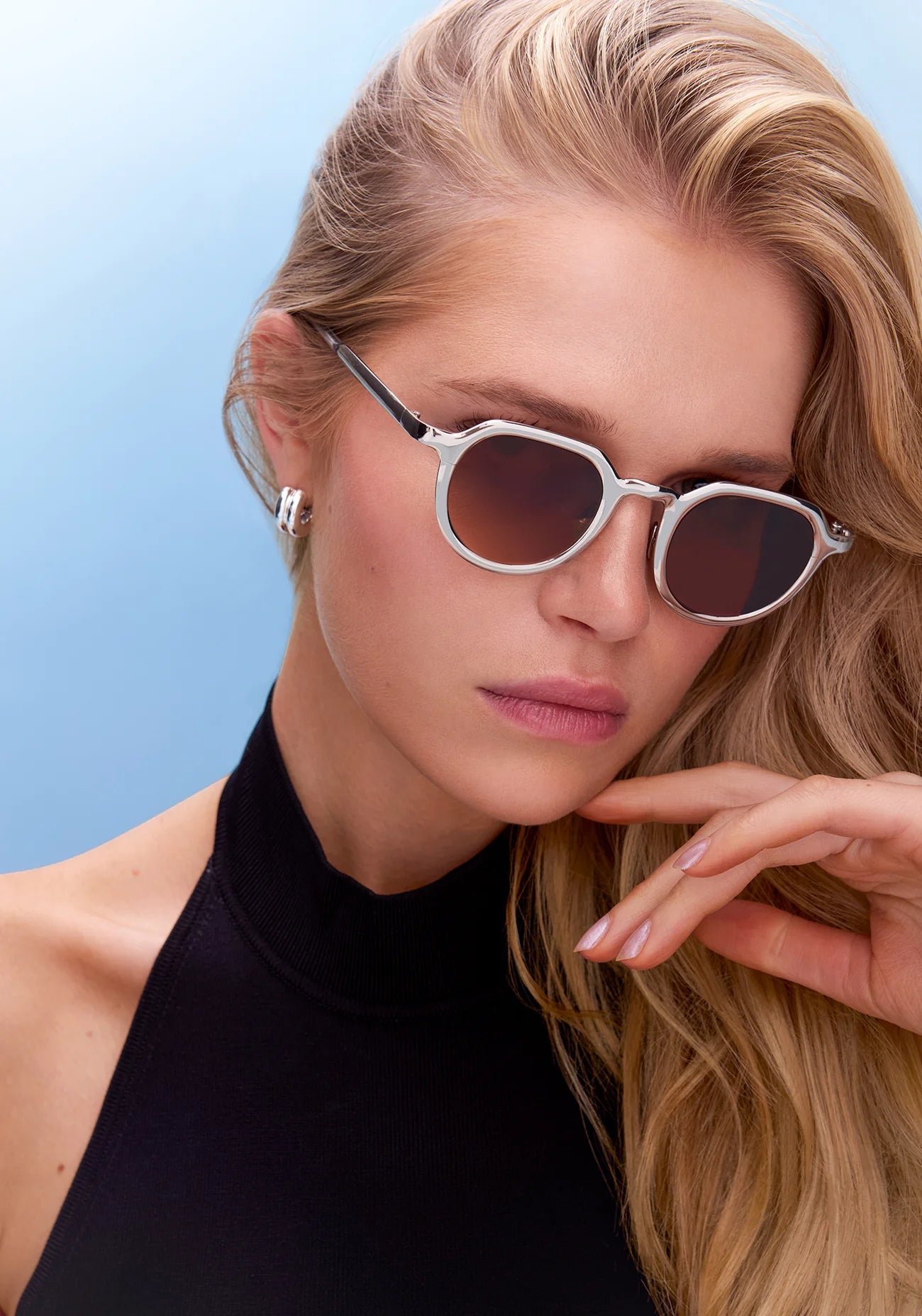 A woman with long blonde hair wears Krewe’s Galvan Sunglasses with an innovative round frame and a sleeveless black top, posing against a light blue background.