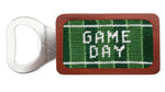 The Smathers & Branson Game Day Bottle Opener features a rectangular wooden handle with green needlepoint and "GAME DAY" in white hand-stitched on it.