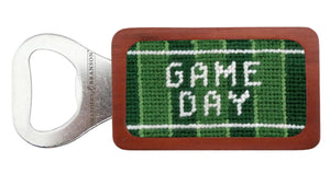 The Smathers & Branson Game Day Bottle Opener features a rectangular wooden handle with green needlepoint and "GAME DAY" in white hand-stitched on it.