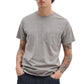 A person wears the Rag & Bone Garment Dye Classic Pocket Tee in light gray, featuring a chest pocket and short sleeves, paired with dark jeans. Tattoos are visible on both arms as they stand with hands in pockets.