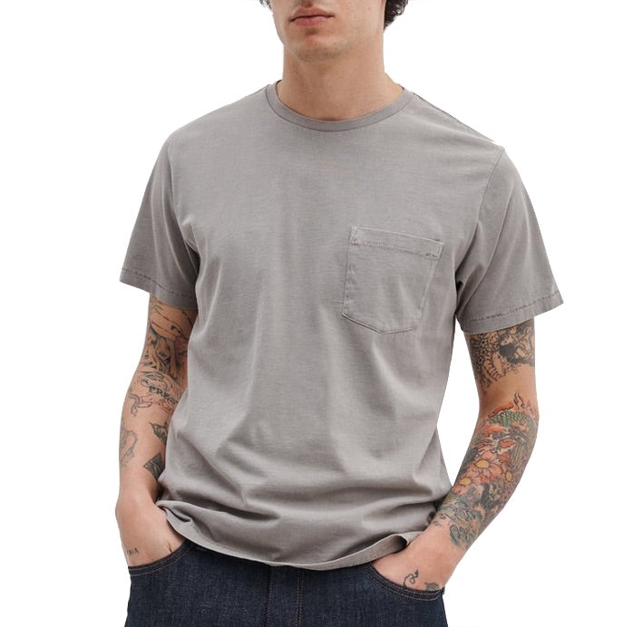 A person wears the Rag & Bone Garment Dye Classic Pocket Tee in light gray, featuring a chest pocket and short sleeves, paired with dark jeans. Tattoos are visible on both arms as they stand with hands in pockets.