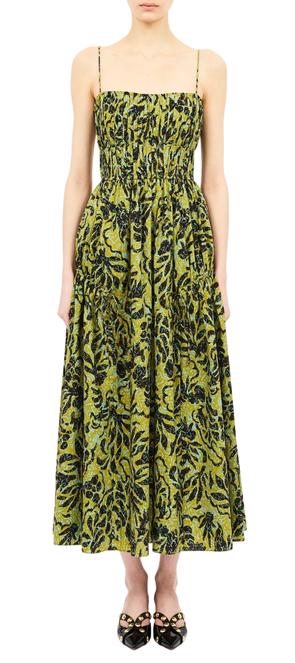 A woman stands against a white background wearing the Ulla Johnson Vidri Cami Cotton Dress—a sleeveless, ankle-length poplin dress with thin straps, a camisole bodice, and a gathered skirt adorned in a green and black leafy pattern—paired with black flat shoes.