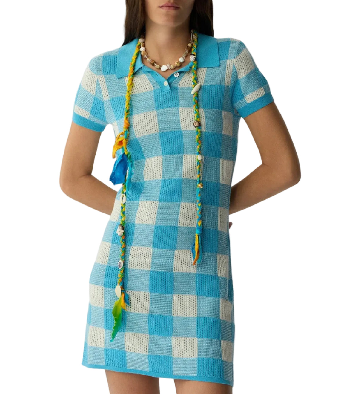 A person wears the Guest in Residence Gingham Polo Dress, featuring blue and white plaid, short sleeves, and a collar, paired with a long beaded necklace decorated in yellow, blue, and green.