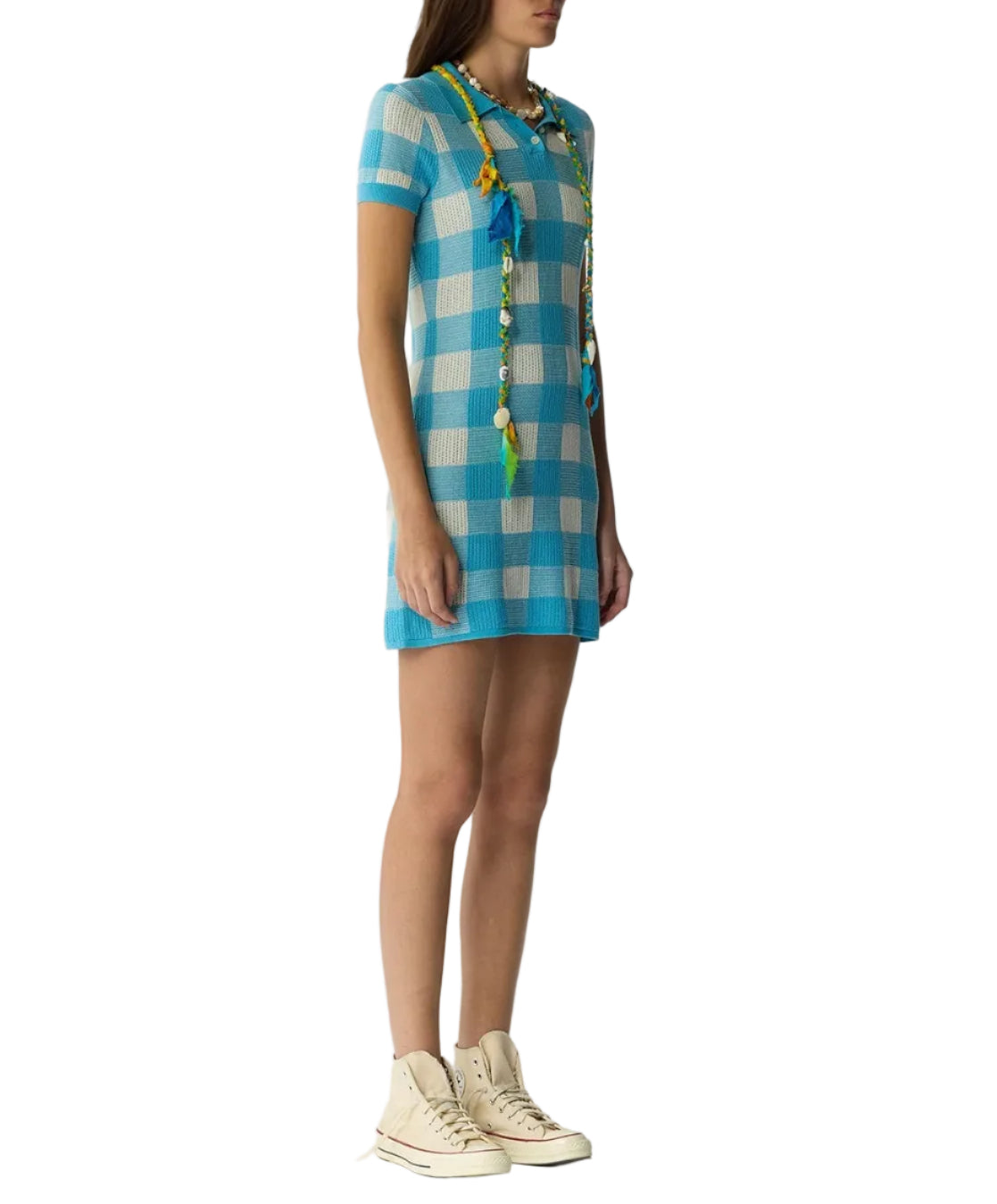 A woman in a blue and white Guest in Residence Gingham Polo Dress, beige high-top sneakers, and a long colorful necklace stands against a plain white background.