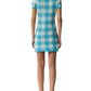 A person stands facing away, wearing the Guest in Residence Gingham Polo Dress in blue and white with short sleeves and beige lace-up shoes.