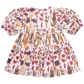 The Pink Chicken Girls' Brooke Dress is a short-sleeved dress adorned with a floral pattern showcasing various flowers in shades of pink, purple, and yellow on a white background. It features gathered puff sleeves and a glitter button back closure that adds an extra touch of charm.