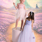 Two girls in pastel dresses, including the Girls’ Goodspell Glimmer Tutu Dress by Tutu du Monde featuring a sparkling sequin bodice, pose before a fantasy backdrop with a yellow brick road, castle, hot air balloons, and flowers. One girl holds a wand; both wear headbands.