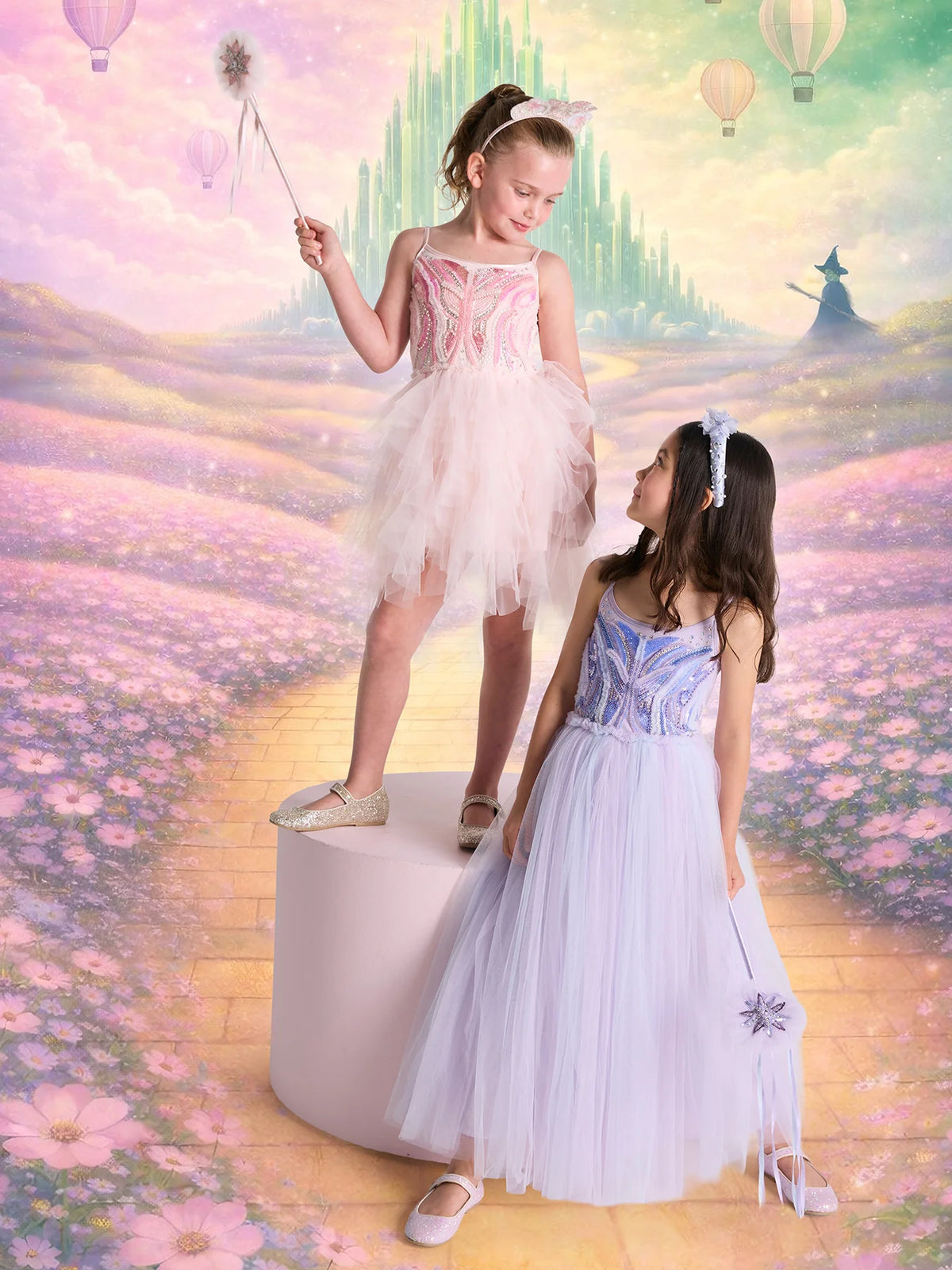 Two girls in pastel fairy dresses, including the sparkling Tutu du Monde Girls’ Glimmering Tutu Dress, stand on a podium against a dreamy backdrop featuring a yellow brick road, castle, hot air balloons, and a distant figure in a hat.