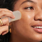 A person uses Supergoop's Destination Glow, a white, travel-friendly facial massage tool, on their cheek, with gold jewelry visible on their hand and ear.