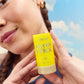 A person holds up a Supergoop Glow Stick SPF 50 sunscreen stick in front of their face against a blue sky, highlighting its portable design for an easy, radiant finish on the go.
