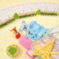 A small blue toy train with a koala figure rides along the track of the Calico Critters Baby Dream Amusement Park playset, while a toy deer stands nearby on a yellow surface adorned with pastel decorations and greenery.