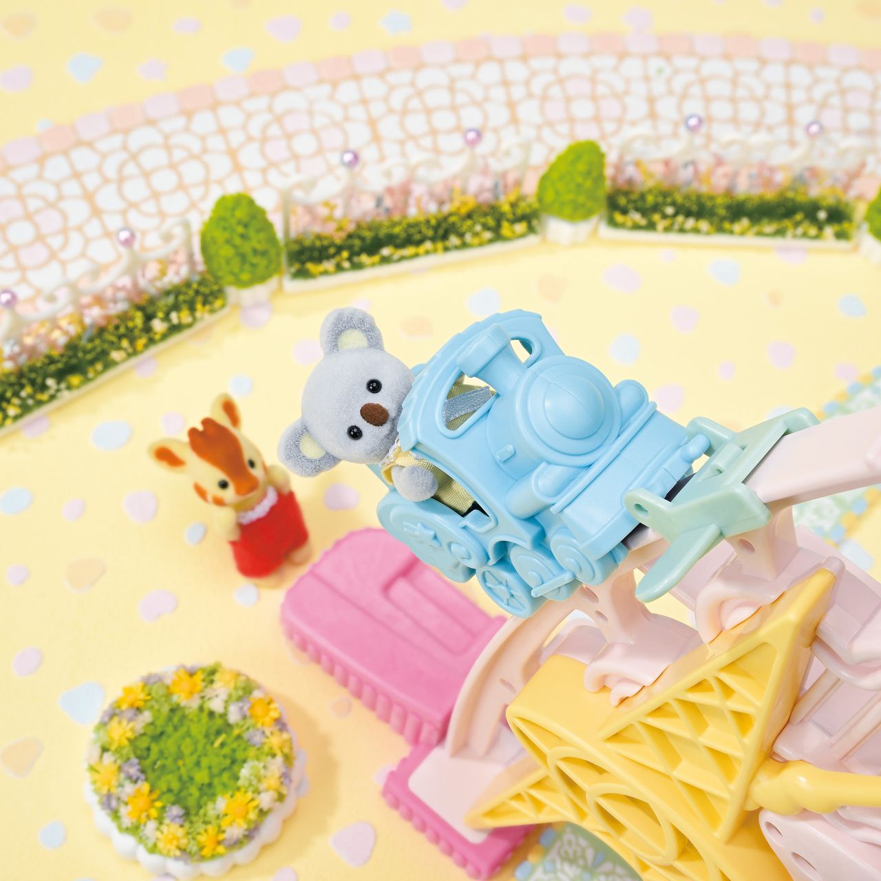 A small blue toy train with a koala figure rides along the track of the Calico Critters Baby Dream Amusement Park playset, while a toy deer stands nearby on a yellow surface adorned with pastel decorations and greenery.
