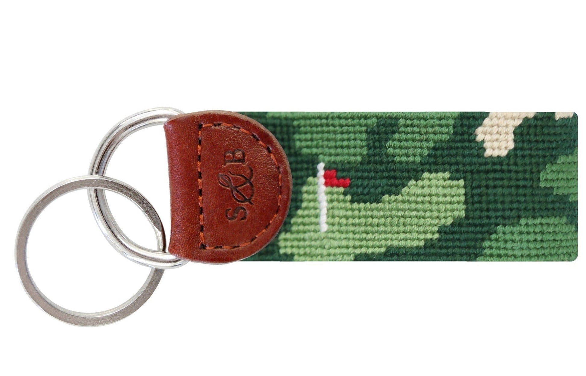 The Smathers & Branson Golfer's Camo Key Fob features an Italian leather tab with two metal rings. Its fabric strap is crafted with French cotton thread, showcasing a green camouflage pattern along with a small red and white flag design.