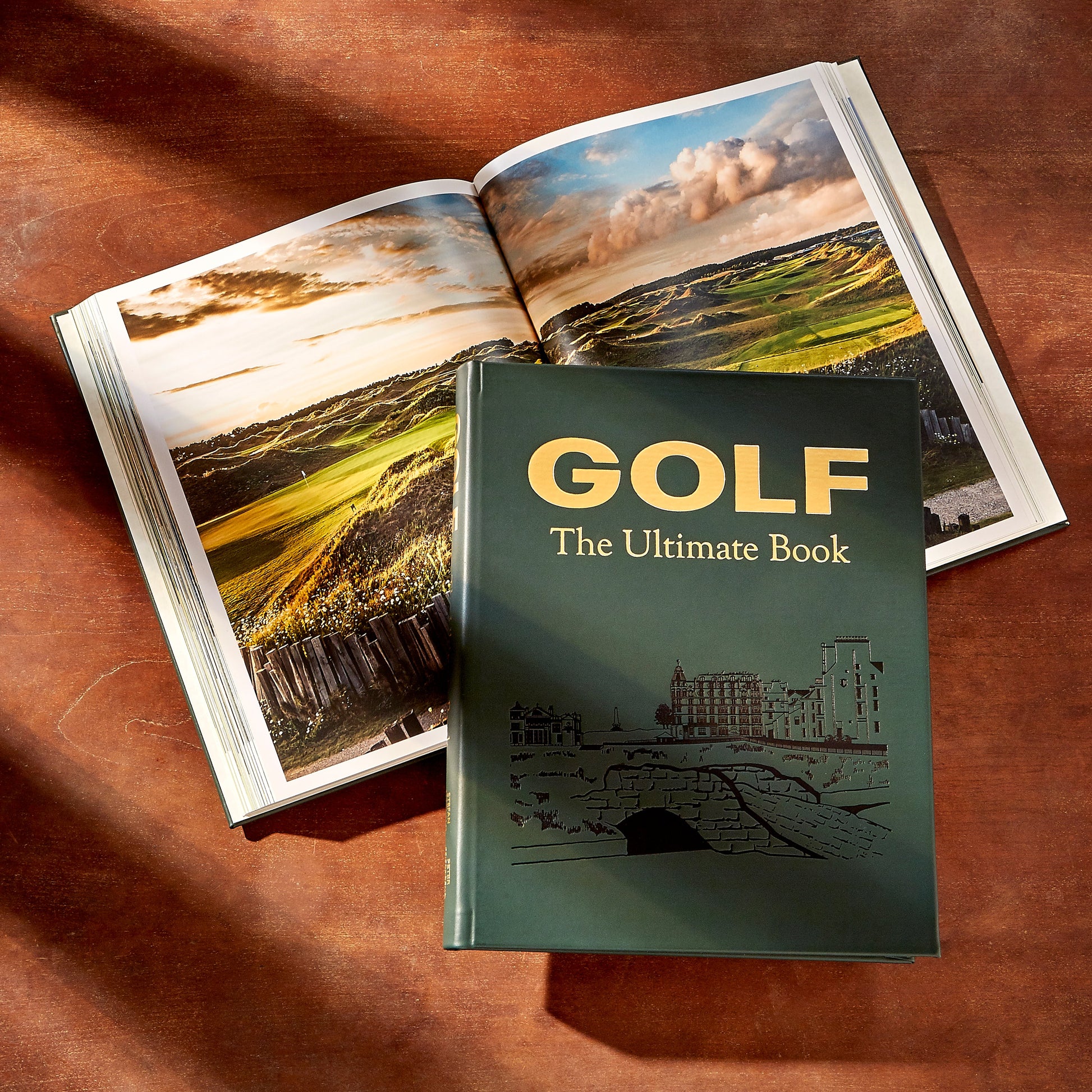 A closed "Golf: The Ultimate Book" by Graphic Image rests on a wooden surface beside an open book showcasing impressive golf courses—an ideal gift for golf lovers.