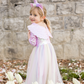 A young child smiles outdoors near white tulips and a stone wall, wearing the Great Pretenders Alicorn Dress with Wings and Headband featuring magical wings and a tulle skirt.