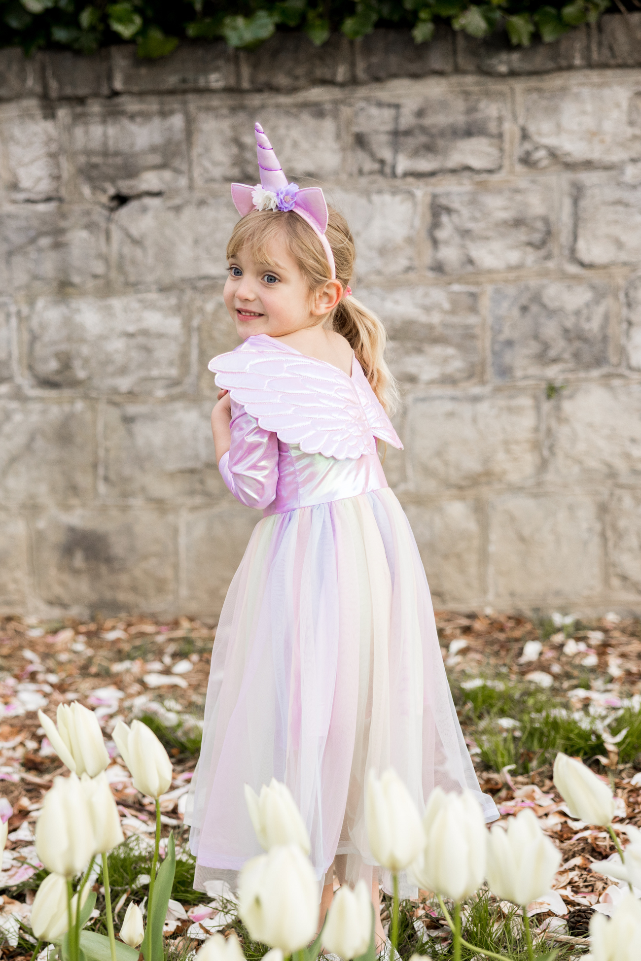 Young girl dressed as a unicorn with Great Pretenders Alicorn Dress with Wings and Headband, smiling over her shoulder in a garden with tulips.