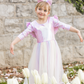 A young girl wears the Great Pretenders Alicorn Dress with Wings and Headband, standing with arms wide in a garden of white tulips, backed by a stone wall and lush greenery.
