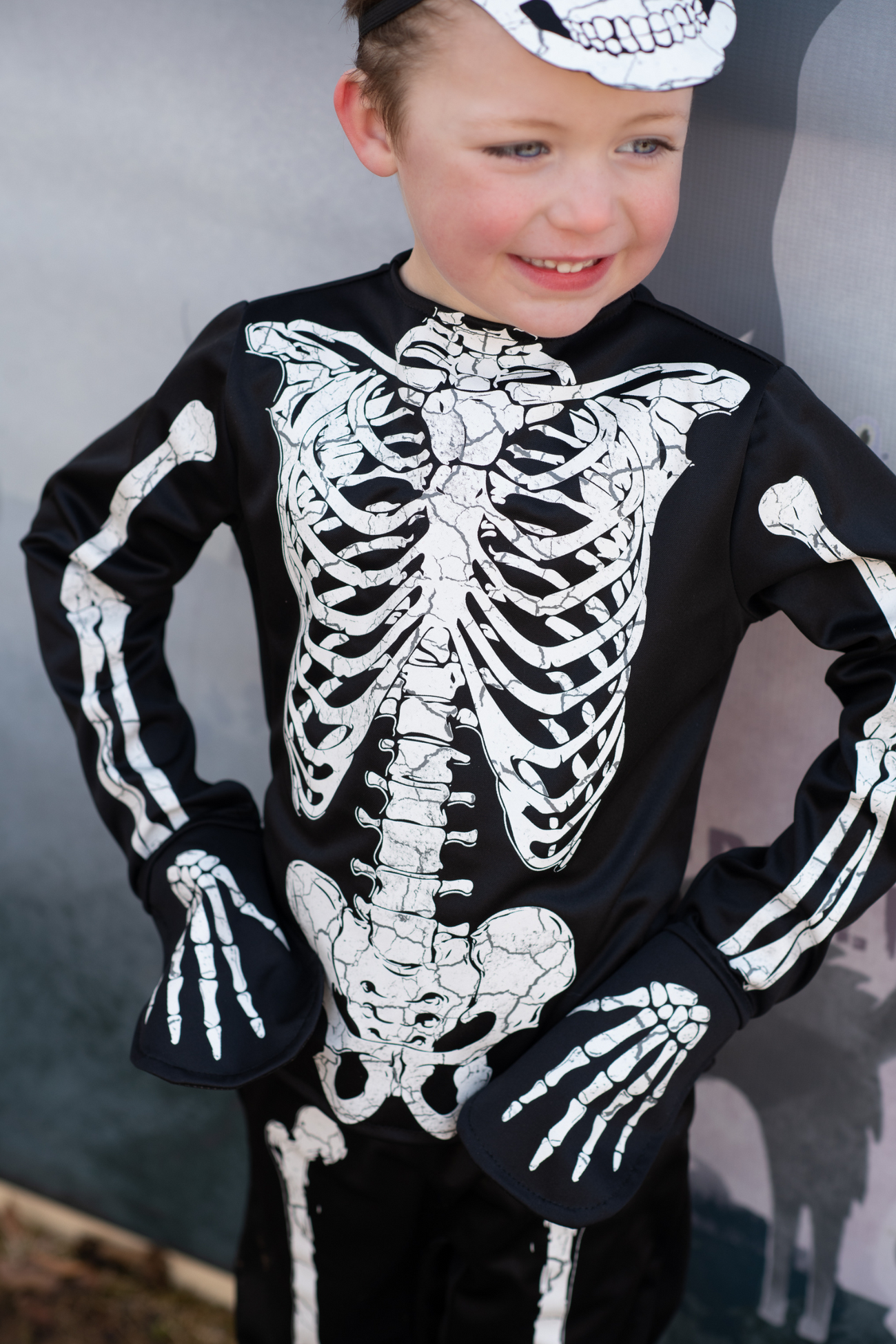 A child dressed in the Great Pretenders Glow in Dark Skeleton Set, featuring white bone patterns on a black costume, stands against a gray background, highlighting its glow-in-the-dark feature ideal for any Halloween celebration.