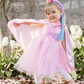 A young child in a pink princess costume with a Great Pretenders Sequins Cape and sequin unicorn headband is posing among blooming tulips and fallen petals.