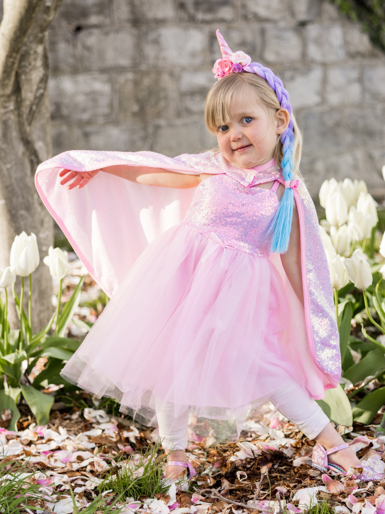 A young child in a pink princess costume with a Great Pretenders Sequins Cape and sequin unicorn headband is posing among blooming tulips and fallen petals.