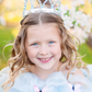 A young princess with a Great Pretenders Cinderella Tiara smiling in front of a blooming tree.