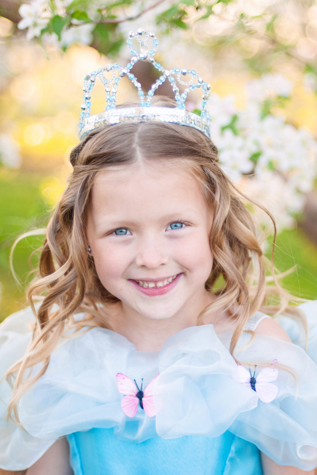 A young princess with a Great Pretenders Cinderella Tiara smiling in front of a blooming tree.