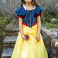 A young girl wearing a Great Pretenders Boutique Princess Gown holds an apple while standing outdoors.