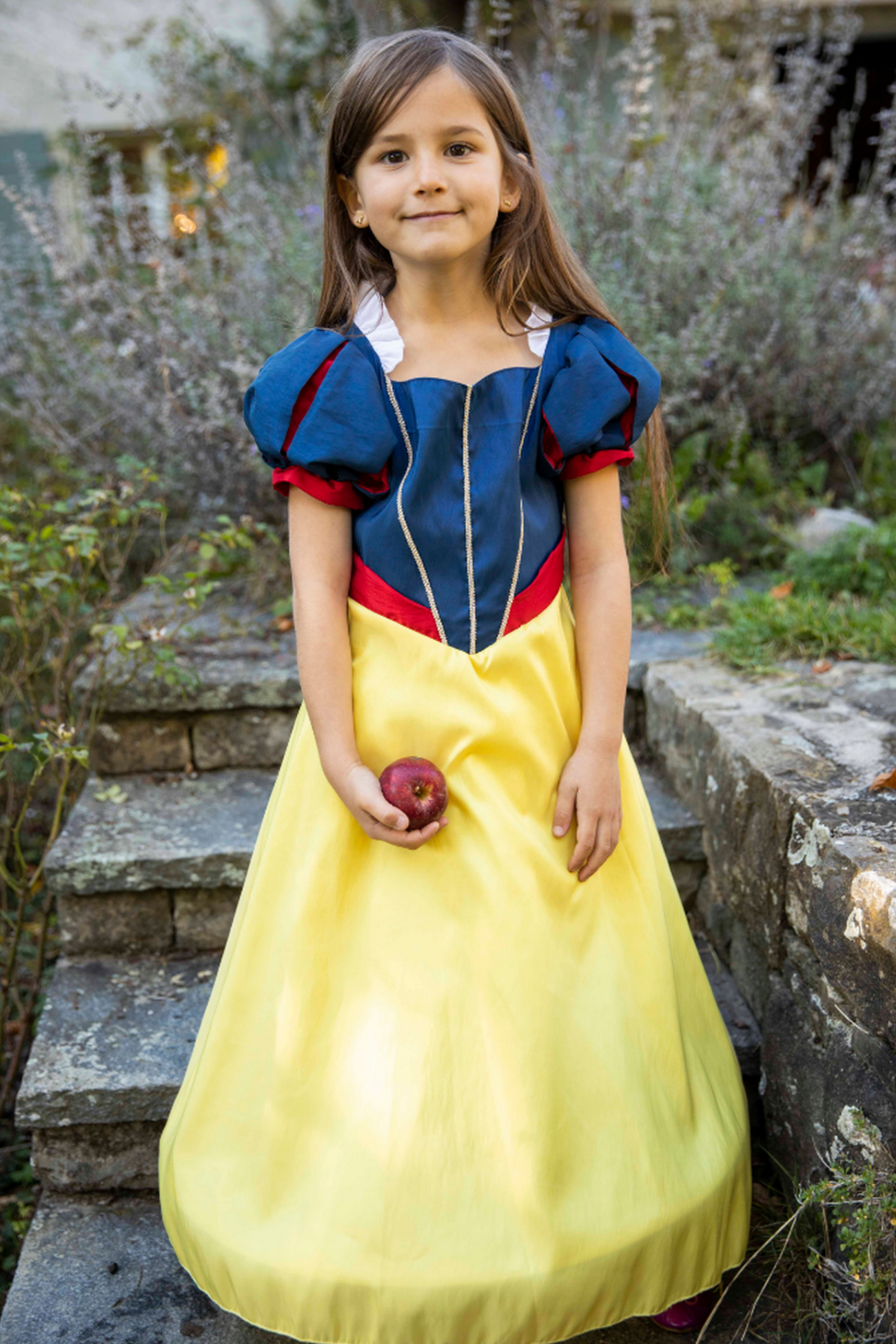 A young girl wearing a Great Pretenders Boutique Princess Gown holds an apple while standing outdoors.