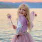 Wearing the Royal Fairy Princess Skirt Set by Great Pretenders, a young child stands by a lake at sunset, looking over her shoulder and smiling, adorned with wings and holding a wand.