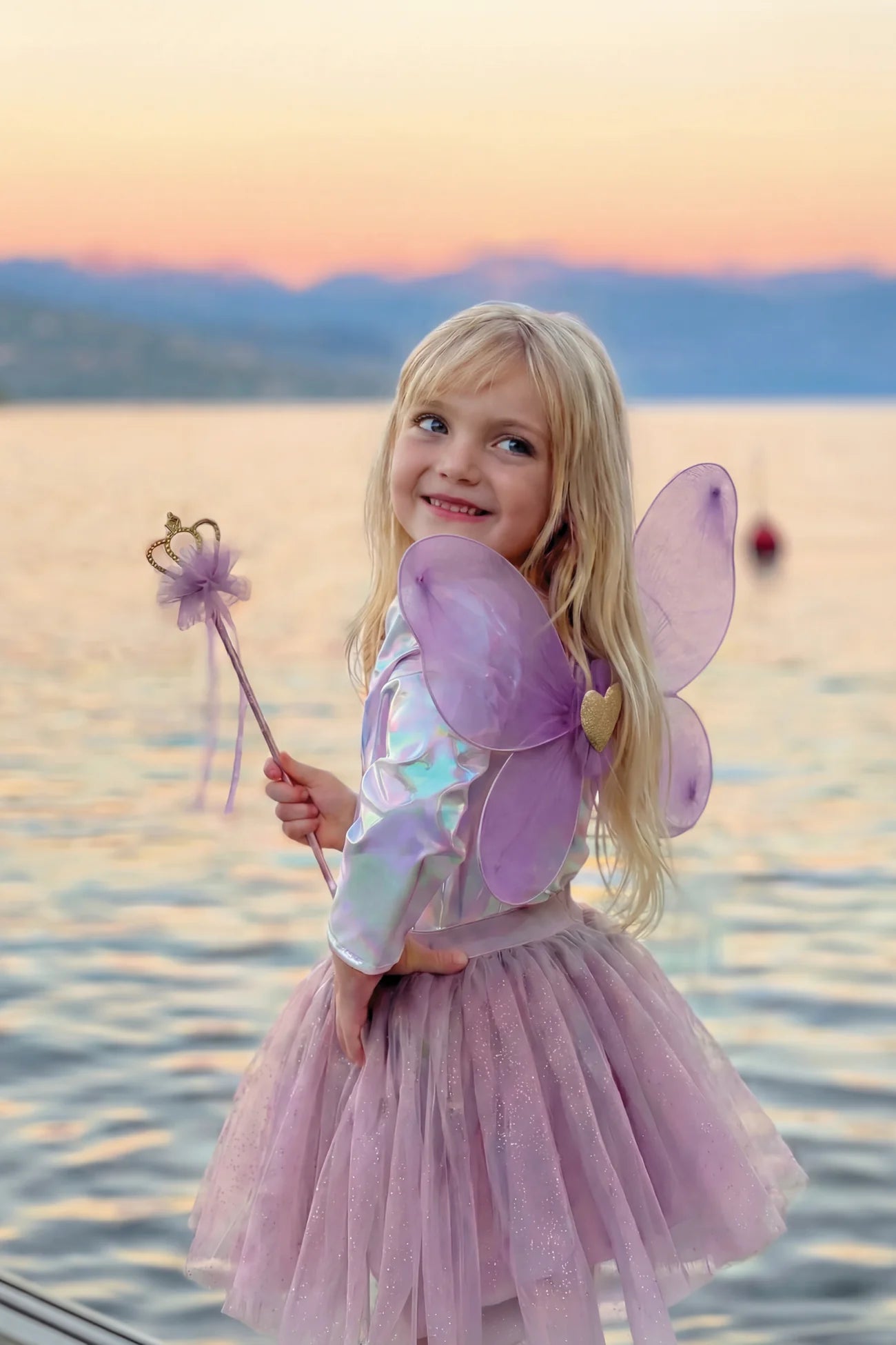 Wearing the Royal Fairy Princess Skirt Set by Great Pretenders, a young child stands by a lake at sunset, looking over her shoulder and smiling, adorned with wings and holding a wand.