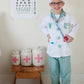 A child, beaming with joy in a Great Pretenders Green Doctor Set with Accessories, stands next to an eye chart and medical jars on a wooden stool, capturing the essence of a dress-up set adventure.