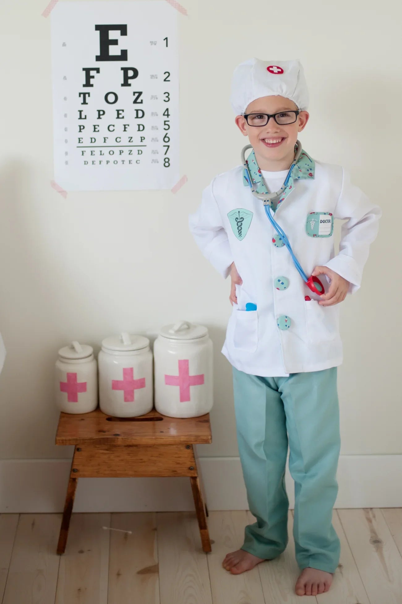 A child, beaming with joy in a Great Pretenders Green Doctor Set with Accessories, stands next to an eye chart and medical jars on a wooden stool, capturing the essence of a dress-up set adventure.