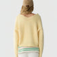 A person facing away wears the Grizzly Double V Sweater by Guest in Residence—a lightweight cashmere pullover in a subtle yellow shade with a double V neckline—styled with a beige cap, white pants, and a green-and-white striped layer visible underneath. The background is plain white.